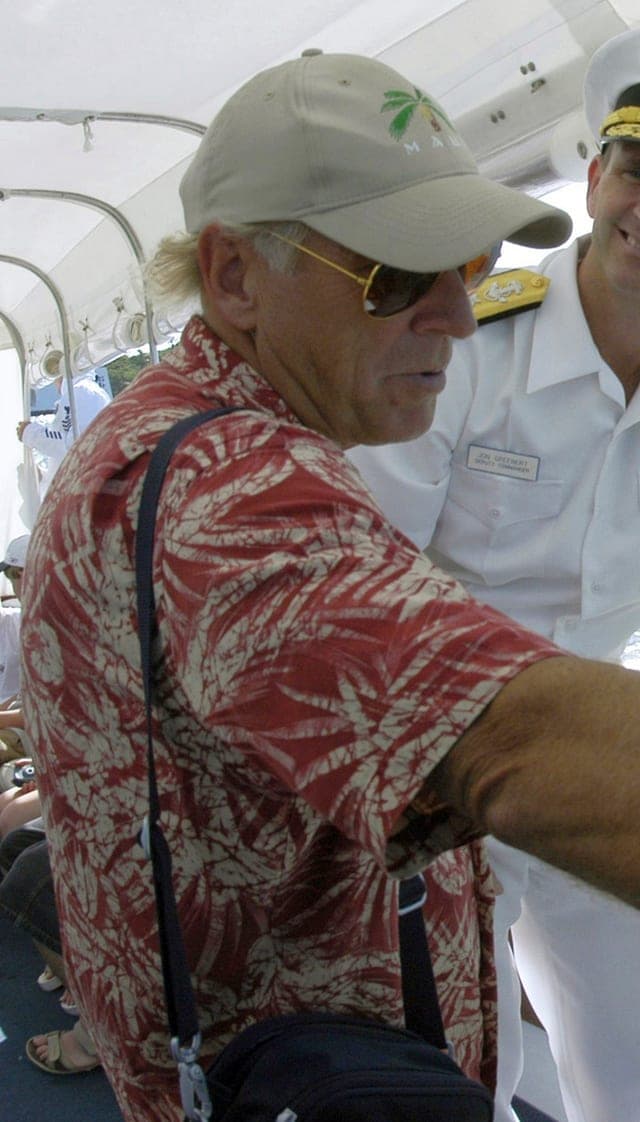 Photograph of the musician Jimmy Buffett in a tropical print shirt and cap aboard a boat.